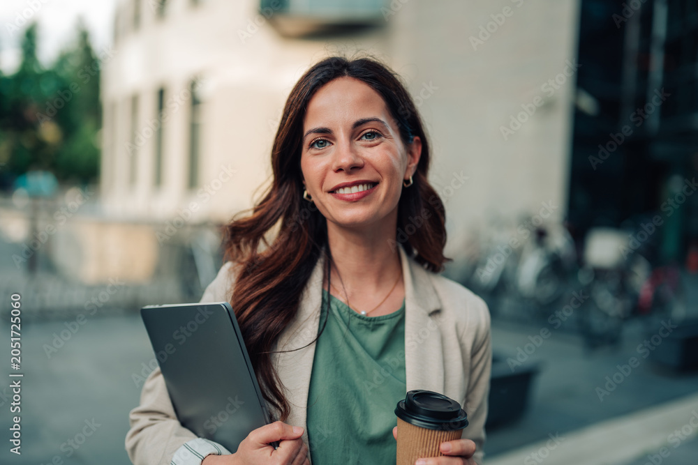 Fototapeta premium Confident businesswoman walking in urban setting holding digital tablet and takeaway coffee cup, smiling at camera