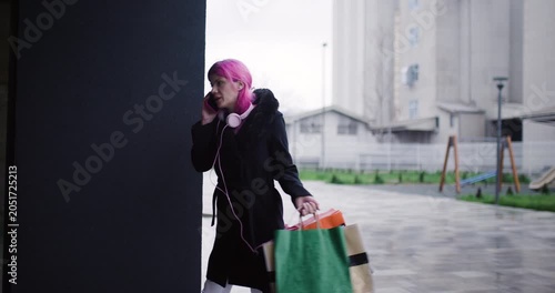Young model with pink hair talking on the phone while carrying shopping bags in urban area during rainy day