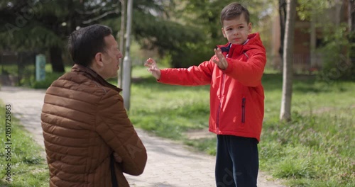 A man and a young boy engage in conversation on a park path surrounded by lush greenery. The sun illuminates the lively atmosphere as they interact thoughtfully