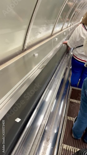 Low-angle shot inside a transit station showing a moving escalator walkway. A person seen from the waist down stands on the right side wearing blue jeans