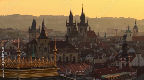 Aerial drone view of the National Theatre gilded rooftop balustrade with Old Town Square towers and Tyn Church silhouetted in warm golden sunrise haze, Prague, Czech Republic.