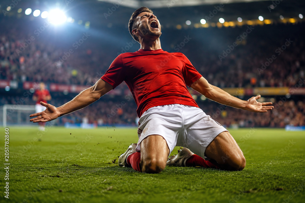 Fototapeta premium Professional soccer player in red jersey celebrating a goal on his knees in a stadium at night. Passionate victory celebration after winning a football match with cheering fans in the background.