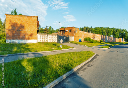 Slika na platnu Sunlit brick industrial compound with perimeter wall and roadside greenery