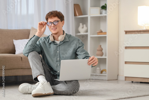 Young man in eyeglasses with laptop sitting on carpet at home