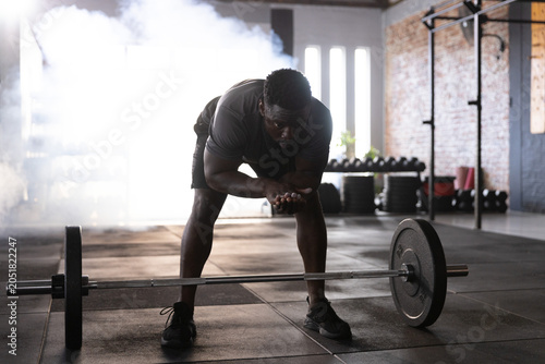 Schilderij op canvas African American man bending over loaded barbell in gym, wearing dark athletic s