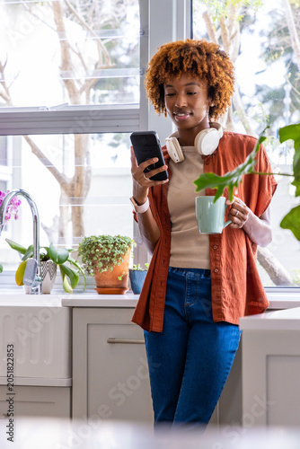 African American adult woman standing by kitchen counter holding mint-green mug checking smartphone