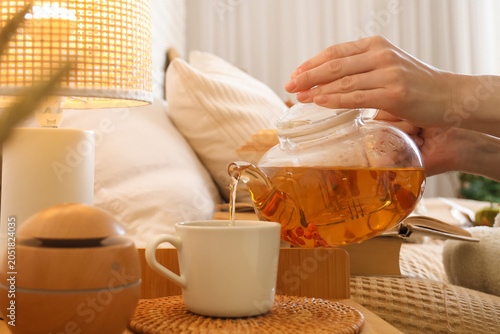 Quadro em tela Female hands pouring green tea from teapot into cup near diffuser and lamp on bedside table in bedroom