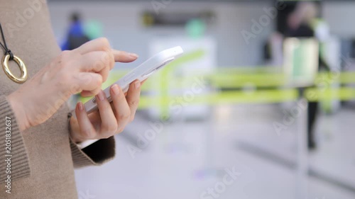 Close-up of woman using smartphone at airport. She focuses intently on her screen. Background shows blurred check-in area with green barriers. Ideal for travel, technology, or communication themes