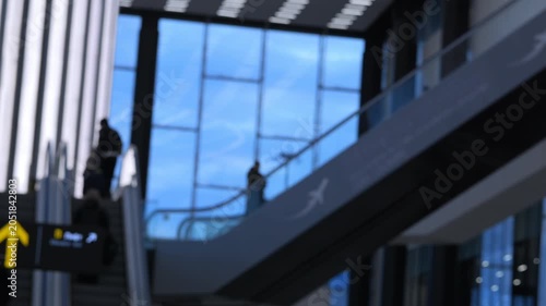 Travelers move on escalators inside modern airport terminal. Silhouettes show people heading toward departure gates. Blue sky visible through large glass windows. Scene conveys busy travel atmosphere