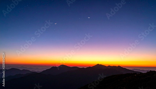 Dawn over the Peaks: The sky transitions from deep twilight blue to fiery orange and yellow as the sun begins to rise over a rugged mountain range, casting silhouetted peaks against the dawn light.
