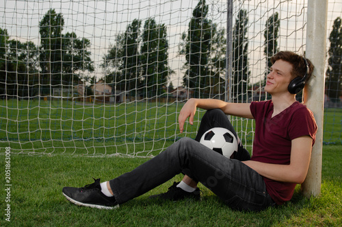 Teenager sitting on the soccer pitch leaned on the goal posing with soccer ball, listening to music