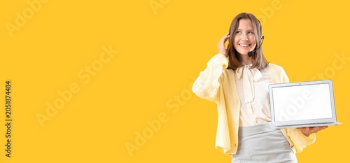 Smiling female call center operator with headset holding laptop on yellow bac...