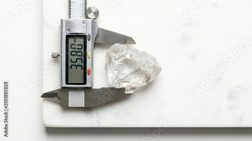 Uncut diamond gemstone accurately measured by digital caliper on white marble slab, representing gemology and jewelry valuation.