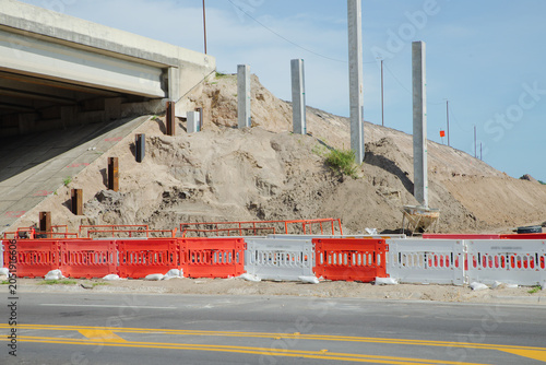 Canvastavla Concrete retaining wall under construction with braced pilings, sandy excavation, heavy equipment and timber shoring along a tree-lined embankment conveying progress and infrastructure development