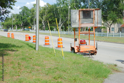 Canvas-taulu An orange portable construction sign trailer and a line of bright traffic cones mark a suburban roadwork zone beside a grassy verge under clear daytime skies