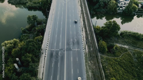Aerial view shows an highway crossing over a river with trees alongside during daylight with rushing cars