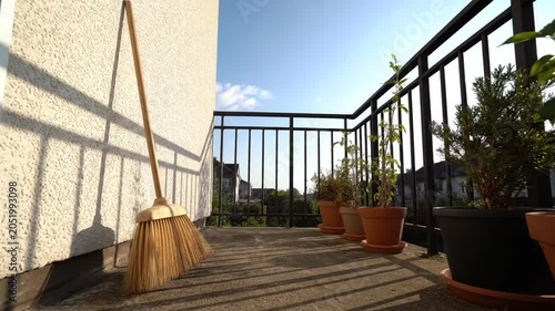 A wooden balcony with a broom leaning against the wall and several potted plants under a clear blue sky.