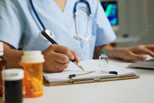 A healthcare professional in scrubs with stethoscope writes notes on a clipboard surrounded by pill bottles and medicine vial, ideal for medical and pharmaceutical content.
