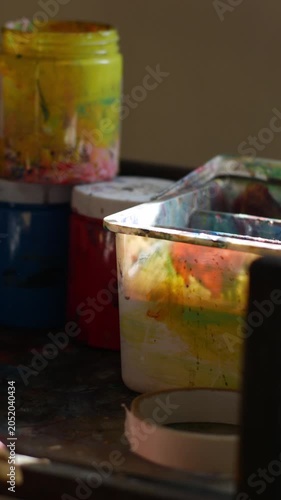 Close-up of single flat paint brush with orange acrylic paint on bristle tip balanced on edge of messy metal palette in dark studio