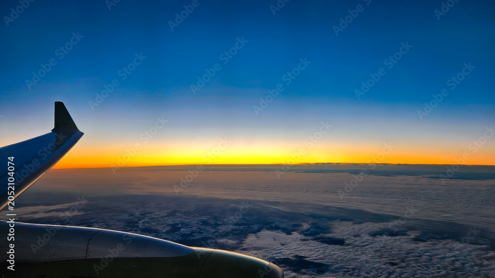 Obraz premium Airplane wing and engine seen from the window, soaring above a fluffy cloud layer, with a colorful gradient horizon and deep blue sky reflecting the beautiful light of dusk or dawn