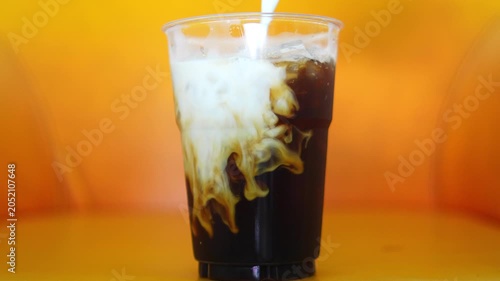 4K Pouring fresh milk into plastic cup of iced black coffee Refreshing iced latte drink isolated on vibrant yellow and orange background.