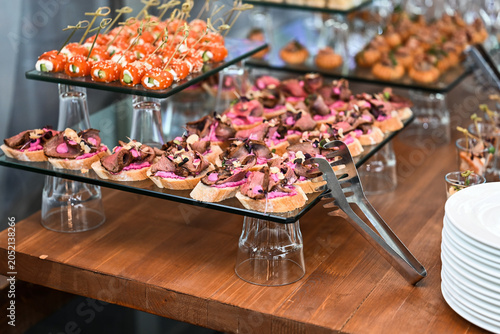 Gourmet buffet table with roast beef bruschetta appetizers on glass stands and metal food serving tongs on wooden counter