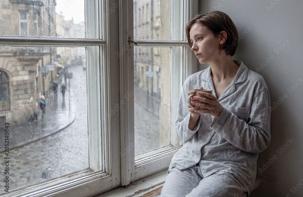 Fototapeta premium A sad girl in pajamas is sitting on the windowsill and drinking tea