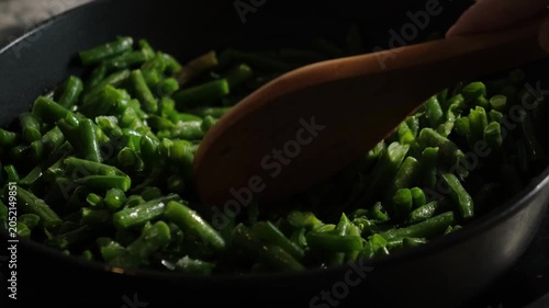 Hand with wooden spoon stirring green bean pods in pan. Preparing plant based vegetarian meal