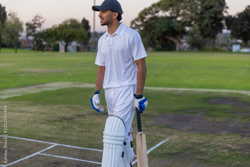 Obraz premium Indian male cricketer standing on pitch at dusk holding bat wearing pads gloves cap