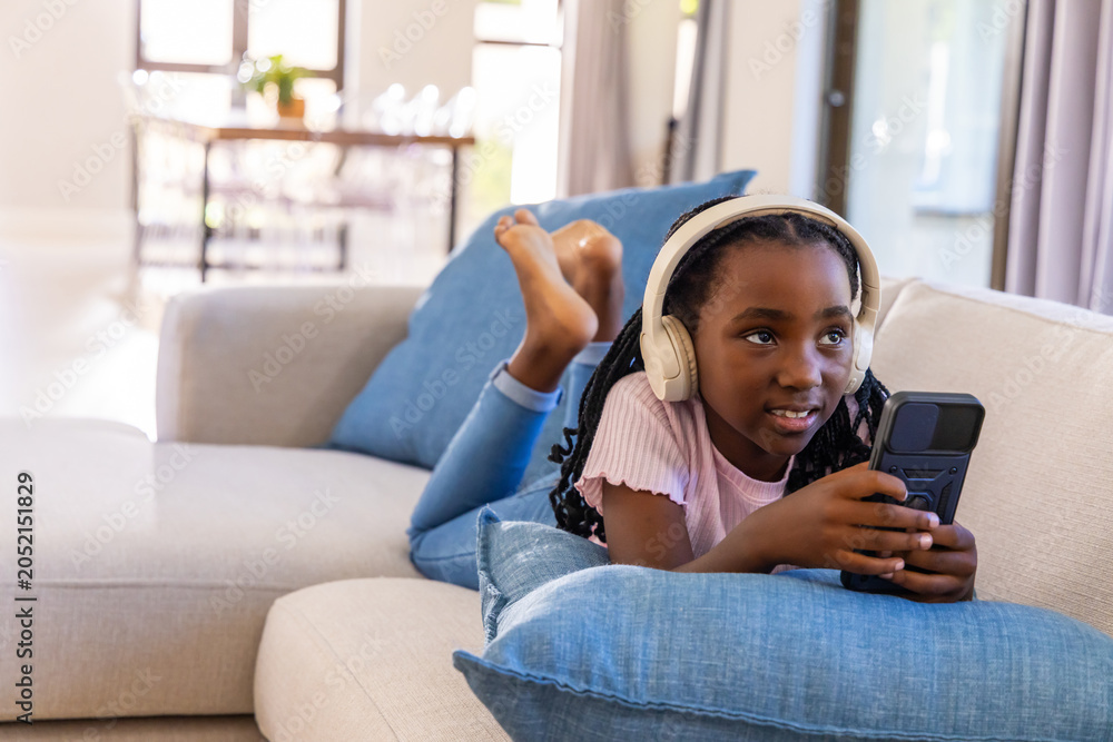 Fototapeta premium School-age African American girl lying on sofa at home wearing white headphones holding device