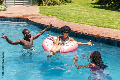 African American friends playing in backyard pool on sunny day with pink donut float
