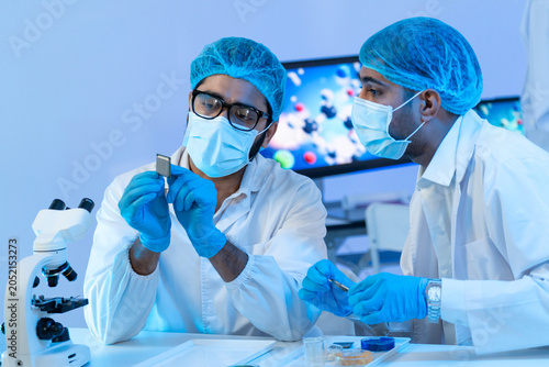 Two male engineers in protective gear carefully examining a microchip semiconductor component together in a high-tech modern laboratory setting.