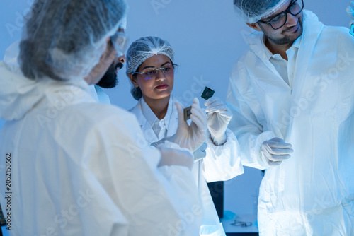 Diverse team of engineers in protective cleanroom suits hold and examine semiconductor microchips together in a modern high-tech laboratory.