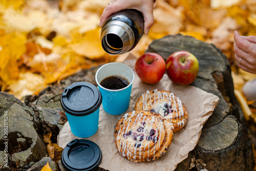 Autumn picnic concept - female hand pouring hot coffee into cup on tree stump with pastries and apples among colorful leaves
