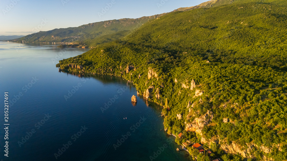 Obraz premium Aerial drone panorama of a scenic lakeside village on Lake Ohrid, North Macedonia. Pristine blue shoreline bordered by lush green mountains and red-roofed houses