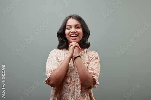 Smiling Indian teenage girl with hands clasped under chin posing against a gray studio background, expressing happiness, confidence and positivity
