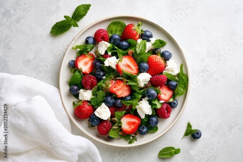 Fresh salad with strawberries, blueberries, raspberries, and goat cheese on a round plate, garnished with mint leaves on a light gray surface