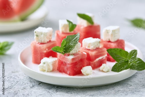 Fresh watermelon cubes topped with feta cheese and mint leaves arranged on a white plate, with a slice of watermelon in the background