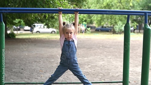 Cute little girl holding onto horizontal bar in park playground, dressed in casual clothes. Girl demonstrates strength and playful energy during outdoor fitness activity in natural setting.