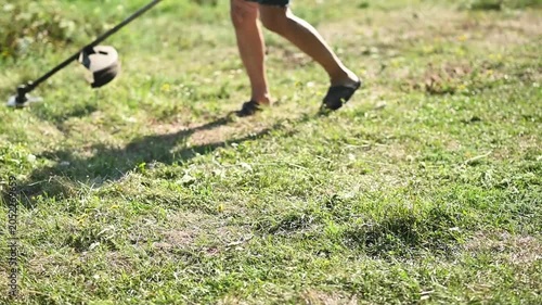 Person using string trimmer to cut tall grass and weeds in sunny garden, closeup of hands operating tool, outdoor landscaping activity, green field and flowers visible, gardening, lawn care concept.