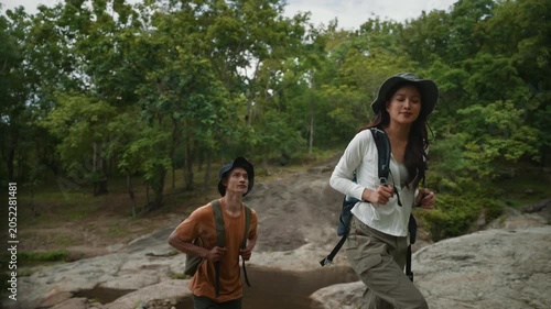 Young Asian couple with backpacks exploring nature together. Man and woman hikers walking on rocks along a stream in a beautiful forest