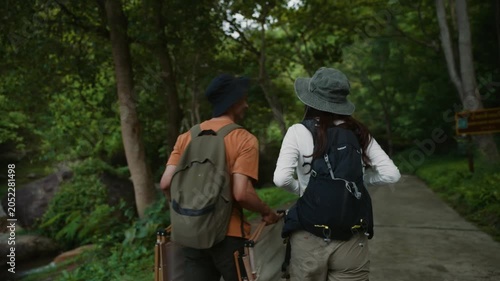 Young Asian couple with backpacks and folding chairs walking on a paved trail through a lush green forest, enjoying their outdoor adventure