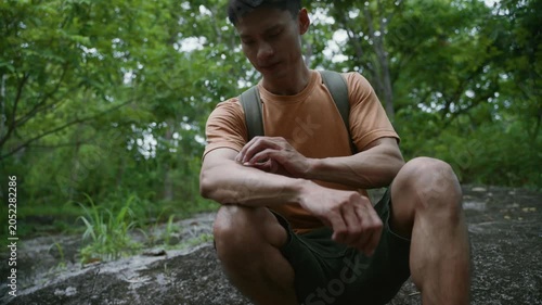 Asian hiker crouching on a rock in the forest while scratching an itchy mosquito bite on his arm