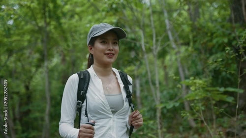 Asian woman with a backpack and hiking poles walking through a lush green forest during the day and checking her phone