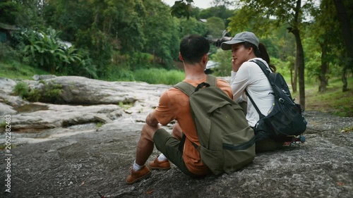 Young adventurous asian couple with backpacks relaxing on a rock and talking during a trekking trip in a green forest