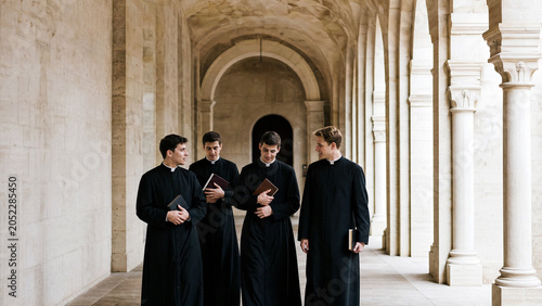 Young seminarians in black cassocks walking stone cloister archway with Bibles, theological study and clergy…