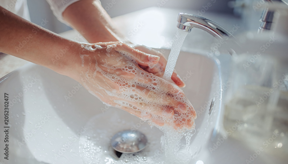 Fototapeta premium A closeup of hands washing with rich soap bubbles under a running faucet in a clean bathroom sink. Perfect for campaigns on personal hygiene, wellness, and health protection.