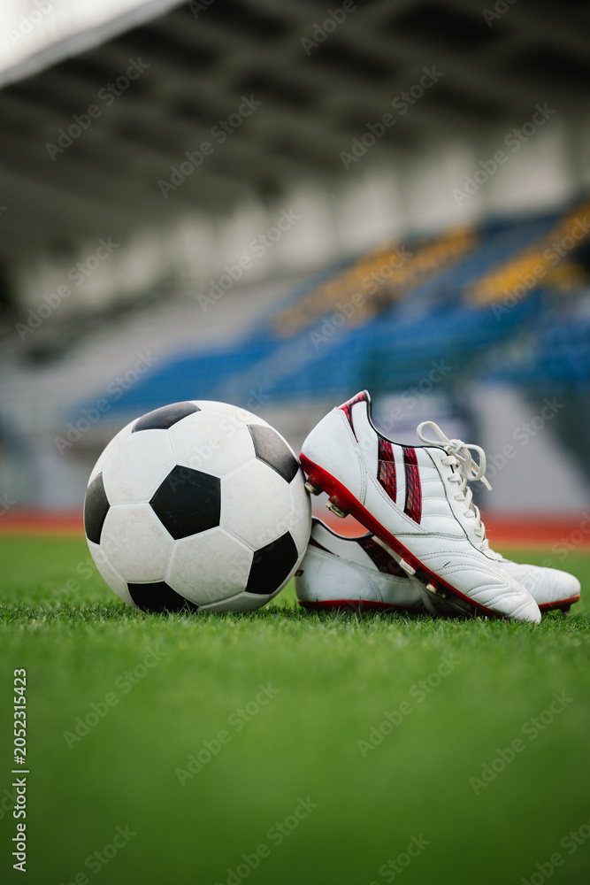 Naklejka premium A soccer ball and football boots rest on a grass field in a stadium, symbolizing teamwork, competition, training, sportsmanship, and the passion of football.
