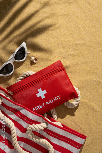Obraz na plátně Red first aid kit on sandy vertical beach next to sunglasses and striped tote fo