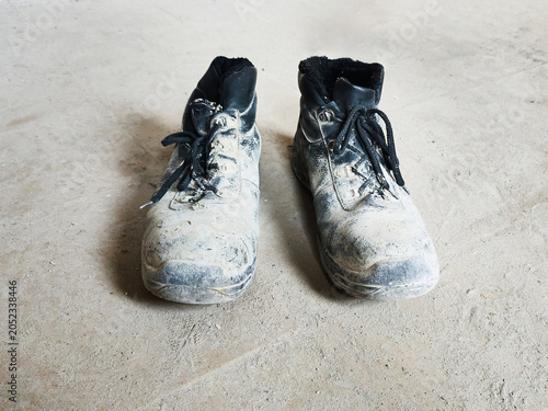 Obraz na plátne A pair of dirty black work boots covered in light-colored dust and mud sits on a plain concrete floor